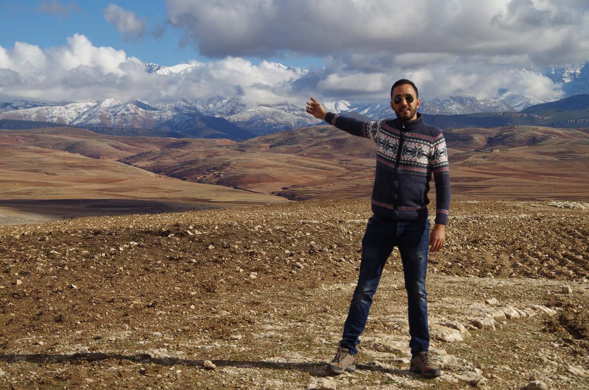 A man gestures across a sweeping rocky landscape below towering mountains and a beautiful sky