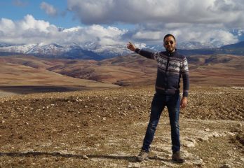 A man gestures across a sweeping rocky landscape below towering mountains and a beautiful sky